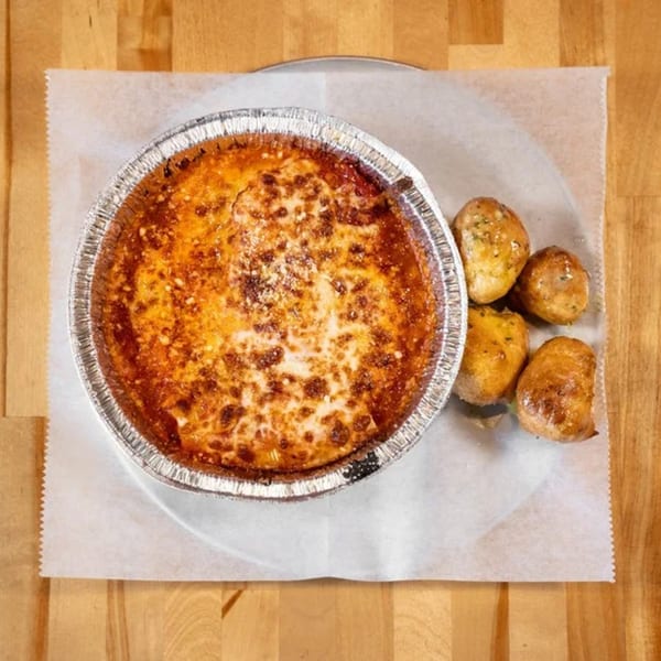 Baked ziti in a foil pan with four garlic knots on parchment paper, on a wooden table.